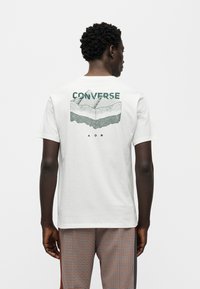 Λευκό βαμβακερό μπλουζάκι με πράσινο γραφικό που απεικονίζει στρωματοποιημένα βουνά και το κείμενο "CONVERSE" στην πλάτη. Κοντά μανίκια, άνετη εφαρμογή.