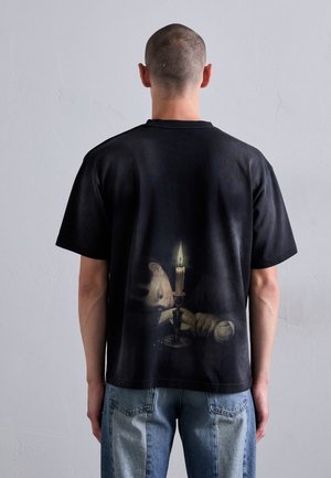 Mand med kort hår iført en sort T-shirt trykt med et stearinlysede scene af en person, der hviler deres hoved ved siden af et tændt stearinlys.
