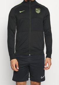 Chaqueta deportiva negra de Nike con cierre de cremallera, logotipo verde y acentos; cuenta con mangas largas y un diseño estructurado, combinada con pantalones cortos negros.