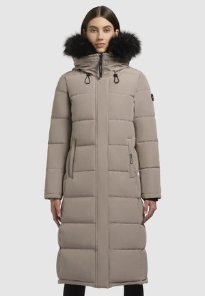Femme portant un long manteau d'hiver matelassé beige avec capuche bordée de fourrure noire, fermeture éclair à l'avant et poches latérales, se tenant contre un fond uni.