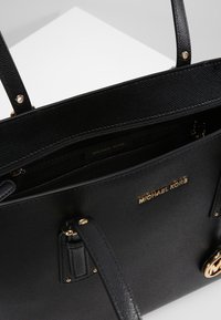 Bolso tote de cuero negro texturizado con herrajes dorados, cierre superior con cremallera, asas dobles y logo de "Michael Kors" en la parte frontal.