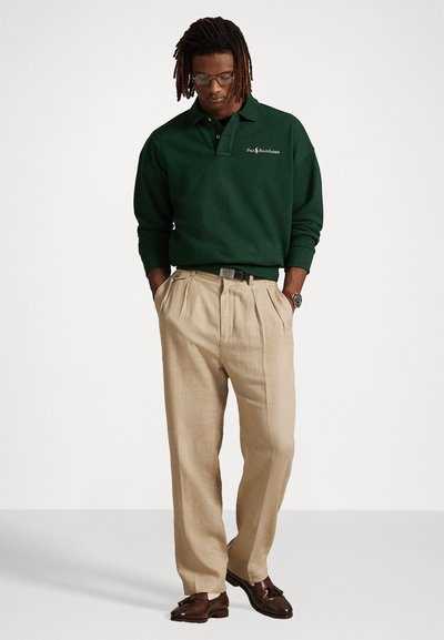 Grønn langermet polo-skjorte med brodert logo, beige folderbukser og brune pensko. Tilbehør inkluderer briller og klokke.