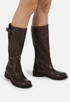 Ein Paar dunkelbraune kniehohe Lederstiefel mit Schnallendetail an den Außenseiten, getragen von einer Person mit nackten Beinen.