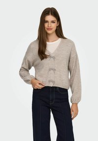 Jasnoszary dzianinowy sweter z dekoltem w serek, trzema dekoracyjnymi kokardkami z przodu i bufiastymi rękawami, połączony z ciemnoniebieskimi dżinsami.