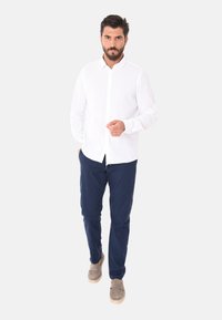 Camicia bianca a maniche lunghe con bottoni, abbinata a pantaloni blu navy e scarpe grigie. Il tessuto sembra leggero e traspirante.
