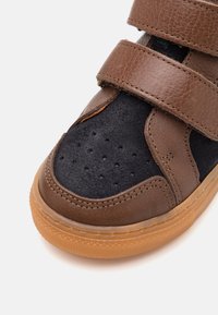 Zbliżenie brązowego i czarnego zamszowego tramwaju z perforacjami i dwiema regulowanymi rzepami, z teksturowaną gumową podeszwą.