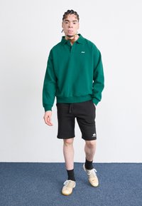 Sweatshirt vert avec un col et un logo, associé à des shorts noirs. La tenue est agrémentée de chaussures de sport dorées et blanches.