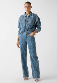 Denim-Outfit mit einer Knopfleiste, ausgefranstem Saum und weiten Hosenbeinen aus einem strukturierten blauen Material mit dezenten weißen Akzenten.