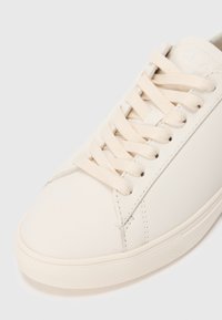 Off-white lace-up sneaker with rubber sole and textured toe cap, angled to show stitching and lace details.