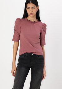 Top scurt roz cu mâneci bufante și decolteu volanat, asociat cu blugi negri. Materialul pare neted și elastic.