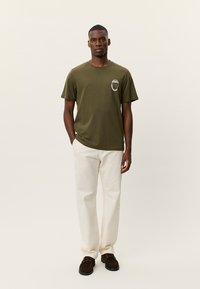 Μπλούζα από βαμβάκι σε ελιά πράσινο με κοντά μανίκια και στρογγυλή λαιμόκοψη, διακοσμημένη με στολισμένο μπαλώμα στο αριστερό στήθος. Συνδυασμένη με παντελόνι κρεμ χρώματος.