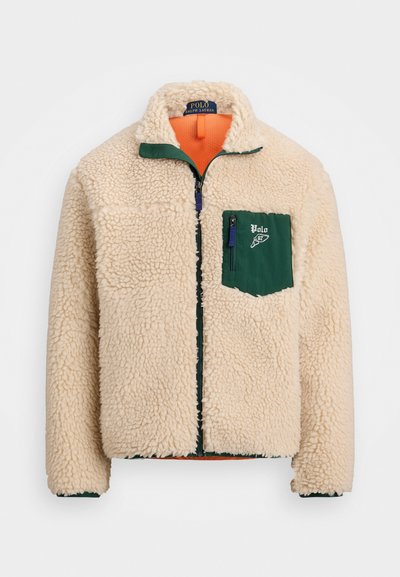 Jaqueta de fleece creme com um bolso com fecho éclair verde e gola, apresentando um forro laranja e fechamento com zíper. Inclui detalhes de costura em contraste.