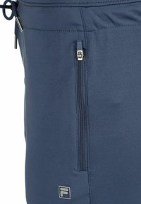 Primer plano de unos pantalones azul marino con un bolsillo vertical con cremallera y un pequeño logo de Fila cerca de la parte inferior del bolsillo.