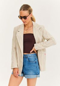 Pruhovaný blejzr v světle béžové barvě s růžovými a modrými akcenty, s klopovým límcem, dlouhými rukávy a kapsou. Spárováno s hnědým topem a džínovými šortkami.