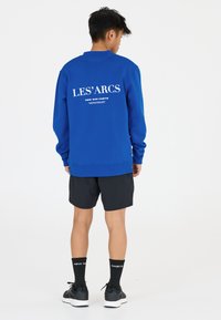 Blauwe sweatshirt met witte tekst op de achterkant, gecombineerd met zwarte shorts en zwarte sokken met witte letters. Casual fit ontwerp.