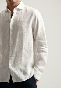 Camisa de lino blanca con cuello abotonado, presenta mangas largas y un sutil brillo. Tela ligera con un corte relajado, con un detalle de marca mínimo.