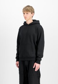 Jonge man met kort blond haar, gekleed in een zwart hoodie en zwarte broek, staand tegen een witte achtergrond.
