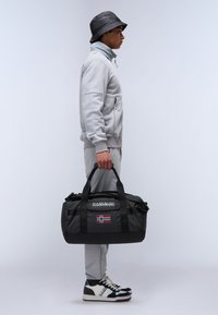 Black duffle bag with logo and Norwegian flag. Smooth, sturdy fabric, dual top handles, and a zippered closure. Worn with grey athletic wear.