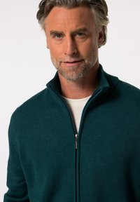 Tyrkysový svetr na zip vyrobený z měkké pleteniny s ribovaným límcem. Zip je stříbrný a svetr má volný střih.