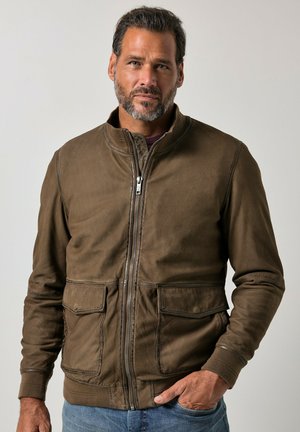 Mann mit kurzen dunklen Haaren und Bart, trägt eine braune Jacke mit Reißverschluss und Taschen sowie blaue Jeans, steht vor einem einfarbigen hellen Hintergrund.