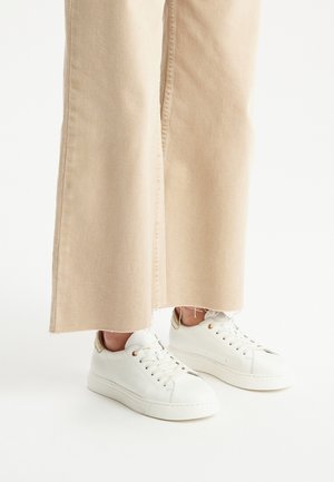 Sneakers bianchi in pelle con superficie testurizzata, dettagli dorati e allacciatura frontale, indossati con pantaloni a gamba larga beige chiaro. Visibile il dettaglio dell'orlo grezzo.
