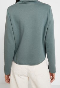Sudadera de tejido suave en un azul claro, con un corte relajado, que presenta un cuello alto y un dobladillo curvado. Combinada con pantalones de pana en color blanco roto.