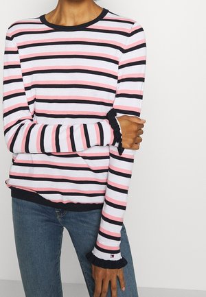 Osoba nosící tričko s dlouhým rukávem s vodorovnými růžovými, černými a bílými pruhy a malými volánky na manžetách, doplněné modrými džínami.