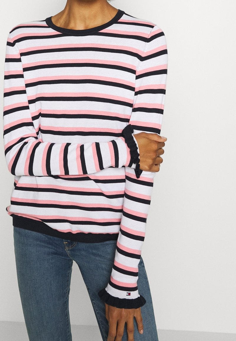 Personne portant une chemise à manches longues à rayures horizontales roses, noires et blanches avec de petites volants aux poignets, assortie à un jean bleu.