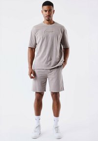 Camiseta de algodón beige con mangas cortas, que presenta un logo texturizado en el frente; combinada con pantalones cortos beige a juego y zapatillas blancas.