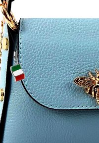 Sac à main en cuir bleu clair avec des détails en or, arborant un emblème de papillon et un petit charme en forme de drapeau italien attaché à la fermeture éclair.