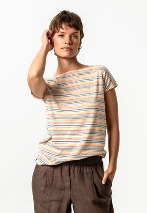 Young person with short hair wearing a multicolored striped short-sleeve shirt and brown pants, hand in pocket and other hand touching hair.