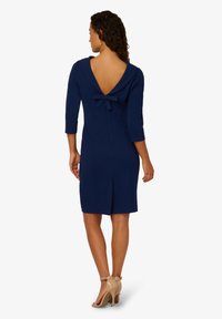 Granatowa sukienka z głębokim dekoltem w kształcie litery V na plecach, rękawami o długości trzech ćwiartek oraz małym detalem w postaci kokardy przy dekolcie; dopasowany krój, do kolan, w teksturowanej tkaninie.
