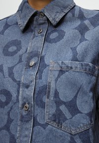 Πουκάμισο από τζιν με μεγάλο λουλουδάτο σχέδιο, μεταλλικά κουμπώματα και τσέπη στο στήθος, όπως φαίνεται από το γιακά έως το μέσο του κορμού.