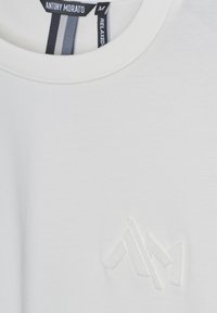 Camiseta de algodón blanca con cuello redondo, textura suave y un logo en relieve en el lado izquierdo del pecho. La etiqueta muestra detalles de la marca y la talla.