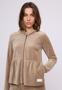 Kapsičková mikina s kapucňou v béžovom zamatovom vzhľade. Má peplum strih, dlhé rukávy a bočné vrecká. Štítok viditeľný na leme.