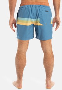 Bañadores de tela azul claro con cinturilla elástica. Presentan rayas diagonales en verde, amarillo y naranja. Incluyen un bolsillo en la parte trasera.