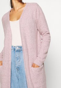 Donna che indossa un lungo cardigan rosa chiaro sopra una canotta bianca infilata in jeans azzurri a vita alta, con le mani nelle tasche del cardigan.
