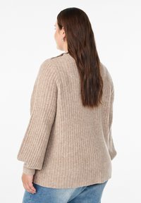 Beige gerippter Pullover mit lockerem Schnitt und glockenförmigen Ärmeln, der eine weiche Textur und einen abgerundeten Saum aufweist, kombiniert mit blauen Jeans.