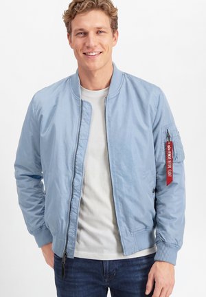 Glunderende man met een lichtblauwe bomberjack over een wit T-shirt en donkere jeans, met handen in de zakken tegen een effen witte achtergrond.