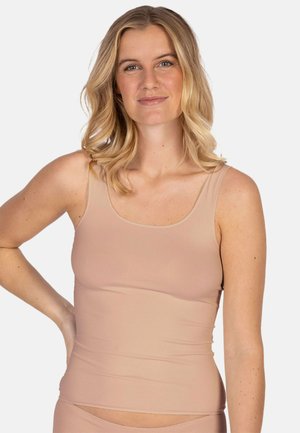 Woman with blonde hair wearing a fitted beige tank top, smiling slightly, with one hand resting on her hip against a plain background.