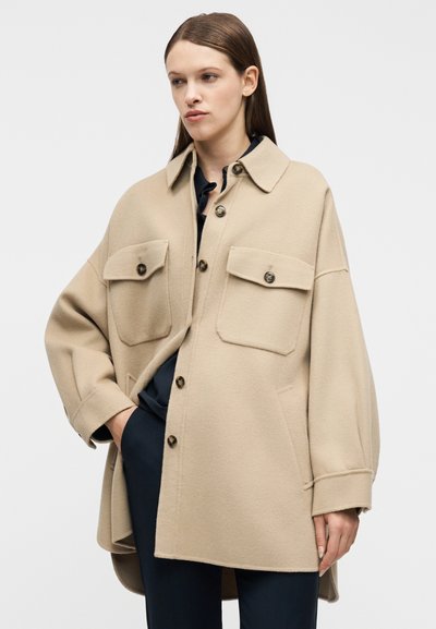 Γυναίκα που φοράει υπερμεγέθη μπεζ παλτό με κουμπιά και μεγάλες μπροστινές τσέπες πάνω από σκούρο μπλε ρούχο, στέκεται μπροστά σε απλό ανοιχτόχρωμο φόντο.