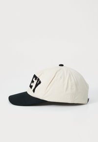 Casquette crème et marine avec une visière courbée, dotée de lettres noires sur le devant et de cinq trous de ventilation. Matériau en coton avec une finition texturée.
