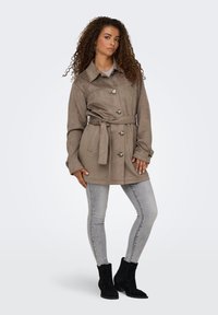 Bruine suède jasje met ceintuur en grote knopen, gecombineerd met lichtgrijze skinny jeans en zwarte enkellaarsjes. Gladde textuur, gestructureerd ontwerp.