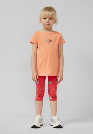 Junges Mädchen mit blondem Haar, das ein pfirsichfarbenes T-Shirt mit Schmetterlingsdruck, rote Leggings mit Schmetterlingsmuster und weiße Turnschuhe trägt, steht vor einem grauen Hintergrund.