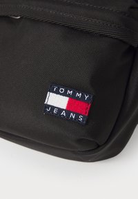 Sac en nylon noir avec un logo Tommy Jeans cousu, accompagné d'accents rouge, blanc et bleu. Surface texturée avec un design épuré.