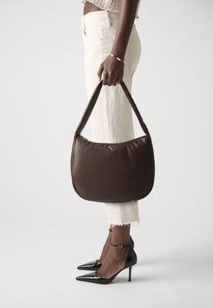 Bruine leren hobo tas met een soepele textuur, voorzien van een gebogen ontwerp en een gouden logo-accent. Gepaard met witte broek tot de enkel en zwarte hakken.