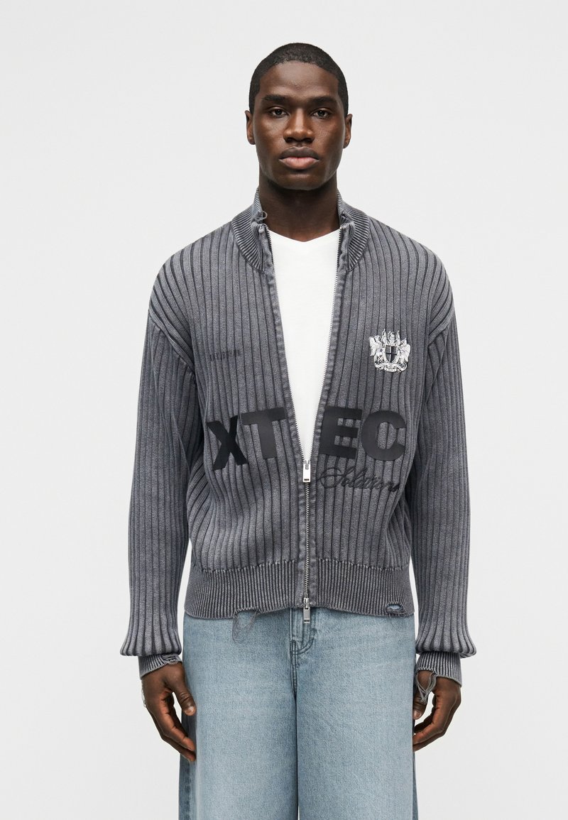 Άνδρας που φοράει γκρι ριμπ ζιπ-απ πουλόβερ πάνω από άσπρο πουκάμισο και ανοιχτό μπλε τζιν, στέκεται μπροστά σε απλό λευκό φόντο.