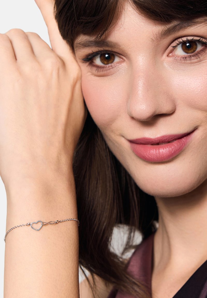Srebrna bransoletka z zawieszką w kształcie serca, o połączonym łańcuszku, noszona na nadgarstku, podkreślająca delikatne detale i finezyjną teksturę.