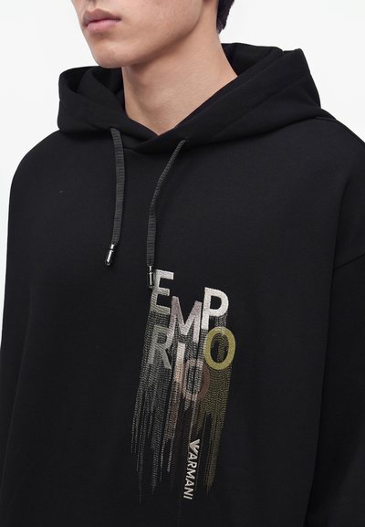 Personne portant un sweat à capuche noir avec cordons et texte brodé "EMPORIO" et "ARMANI" en couleurs mélangées sur la poitrine.