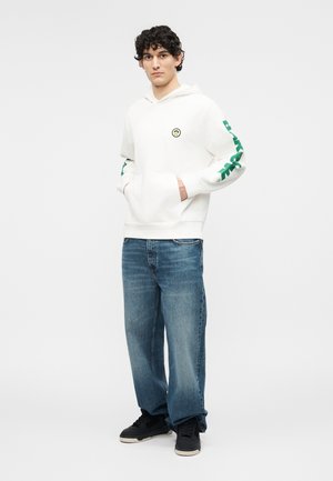 Jeune homme portant un sweat à capuche blanc avec des lettres vertes sur les manches, un jean bleu et des baskets noires, debout les mains dans les poches du sweat.